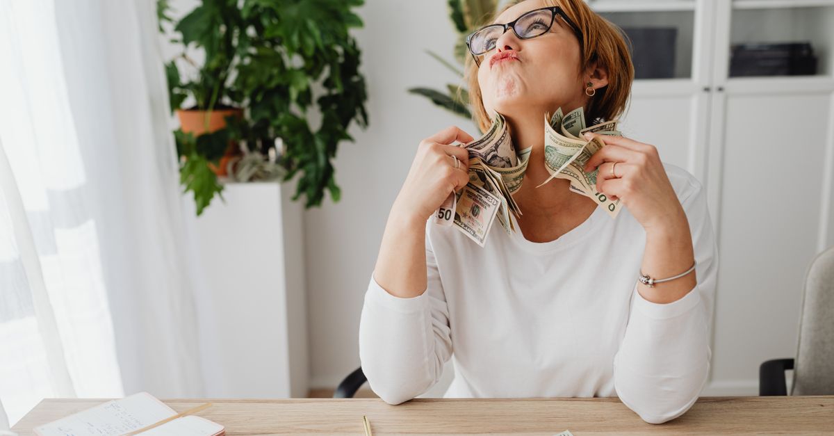 A happy woman holding dollar bills, sitting at a desk with plants and office supplies.