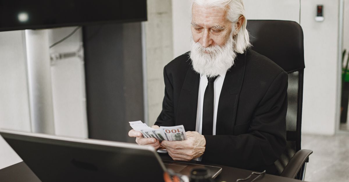 Senior businessman in suit counting money at office desk with a laptop. Elegance and financial focus in modern workspace.