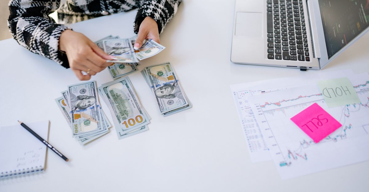 Person counting cash next to laptop and stock market charts on a white table.