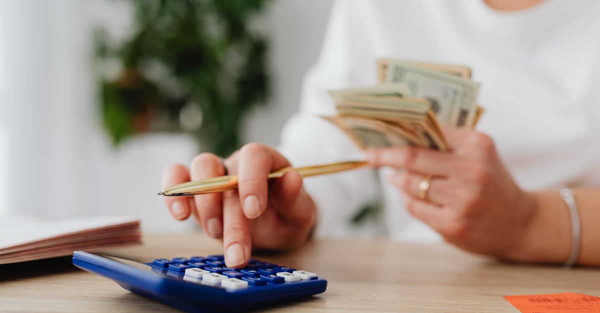 A person sits at a desk calculating finances using a calculator and holding cash.